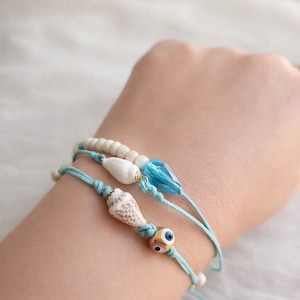 May include: Three handmade bracelets with a beach theme. The bracelets feature light blue cords, seashells, white beads, and a blue crystal. One bracelet has a gold-colored bead with an evil eye design.