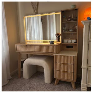 May include: A light wood vanity with a large, illuminated mirror. The vanity has a built-in shelf with glass doors, three drawers, and a matching stool. The mirror is surrounded by a warm, glowing light. The drawers have gold-colored handles.