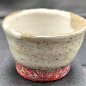 May include: A small ceramic cup with a speckled off-white upper section and a vibrant red base featuring a detailed, intricate floral and leaf pattern. The rim is uneven and thick, showing a natural, hand-crafted texture with visible clay flecks.