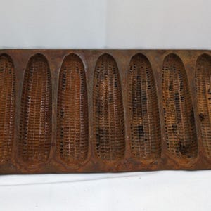 May include: A vintage cast iron cornbread pan with seven corn cob molds. The pan is rectangular with rounded ends and handles. The surface has a rustic, aged appearance with a brown and orange color.