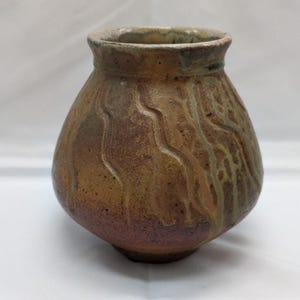 Puede incluir: Un pequeño jarrón de cerámica bulboso con un borde ancho y acampanado. El exterior presenta un esmalte terroso y texturizado en tonos marrón, bronceado y verde apagado, con crestas verticales onduladas en relieve y una base oscura y moteada.