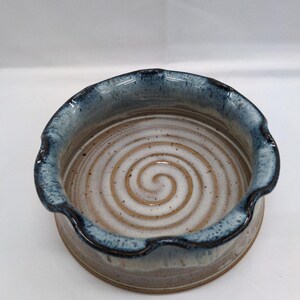 May include: A handmade ceramic bowl with a spiral design in the center. The bowl has a wavy, dark blue rim and a light brown and white interior. The exterior is a light brown color.