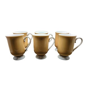 Puede incluir: Un juego de cinco tazas a juego con un exterior liso de color naranja claro y un interior blanco. Cada taza cuenta con un asa curva y una base de pedestal distintiva, lo que crea una apariencia uniforme en toda la vajilla.