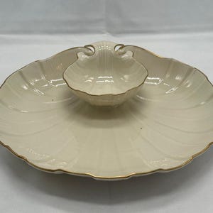 May include: A cream-coloured ceramic serving dish shaped like a large scallop shell with a smaller, integrated shell-shaped bowl in the centre. The edges of both the large platter and the central bowl are trimmed with a thin, metallic gold line.