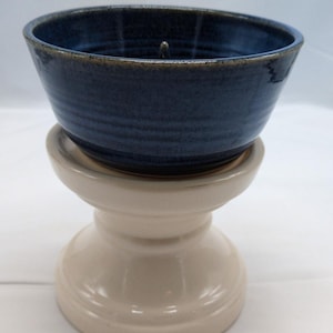 Puede incluir: Un portavelas de cerámica con una parte superior en forma de cuenco azul oscuro y una base de pedestal beige. El cuenco tiene una superficie texturizada y una pequeña pieza de metal en el centro. El portavelas está diseñado para sostener una vela.