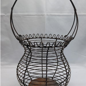May include: An antique wire basket with a handle. The basket has a rounded body and a scalloped rim. The metal is a dark, aged brown color. The basket is designed to hold items and has a rustic, vintage aesthetic.