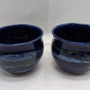 Puede incluir: Dos cuencos de sopa de cerámica hechos a mano. Los cuencos son de color azul marino oscuro con un acabado brillante. Cada cuenco tiene una forma redondeada con un asa y una banda decorativa verde y blanca cerca de la base. Los cuencos tienen unos 15 cm de diámetro.