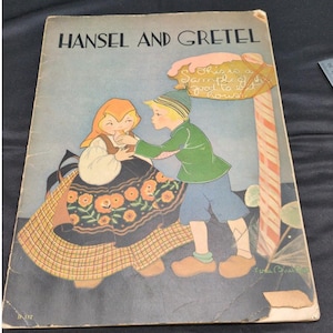 Könnte beinhalten: Vintage Kinderbuchcover mit dem Titel "Hänsel und Gretel". Die Illustration zeigt zwei Kinder in traditioneller Kleidung. Das Mädchen trägt eine orangefarbene Kopfbedeckung und einen geblümten Rock. Der Junge trägt eine grüne Jacke und blaue Hosen. Ein Schild lautet: "This is a sample of good to eat houses."
