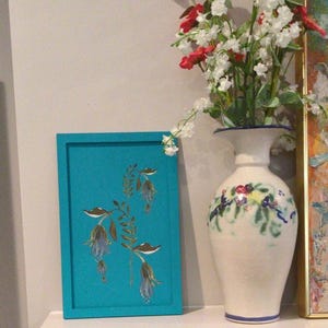 Op de afbeelding: Een ingelijst turquoise kunstwerk met een bloemenontwerp met blauwe en gouden accenten. Daarnaast staat een witte vaas met bloemmotieven met rode en witte bloemen. Het kunstwerk en de vaas staan op een witte plank.