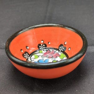 Puede incluir: Un pequeño cuenco de cerámica redondo con un exterior naranja vibrante y un borde negro. El interior presenta una base blanca decorada con un patrón floral central en tonos rosa, azul y verde, rodeado de detalles de puntos negros y blancos.