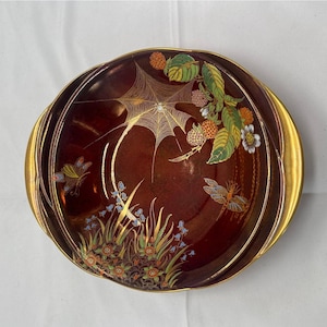 Peut inclure: Un bol en céramique rond et profond, brun foncé, avec des anses dorées et un bord peint en or. L'intérieur présente une toile d'araignée dorée détaillée, une libellule, un papillon, des feuilles vertes, des baies orange et des fleurs bleues.