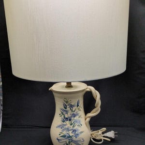 May include: Cream ceramic lamp with a white lampshade. The lamp base is shaped like a jug with a braided handle and blue floral design. The lamp has a standard electrical plug.