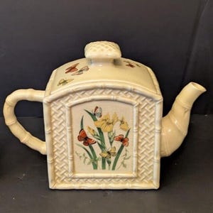 Può includere: Un servizio da tè color crema con una teiera quadrata e due tazze abbinate. La teiera ha un manico e un beccuccio in stile bambù, con un coperchio. Ogni pezzo è decorato con un motivo floreale e farfalle all'interno di una cornice rettangolare.