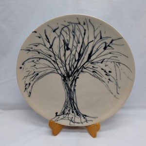 May include: A round, off-white ceramic plate featuring a hand-drawn tree design in dark blue. The tree's branches and roots are depicted with expressive lines and dots. The plate is displayed on a small, gold-colored stand.