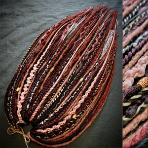 Puede incluir: Un conjunto de rastas hechas a mano en tonos burdeos, marrón, negro y rosa. Las rastas presentan varias texturas, incluyendo trenzas, giros y secciones envueltas, con cuentas doradas y detalles de encaje blanco. Las rastas están atadas con hilo natural.
