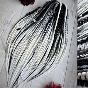 May include: A collection of black, white, and grey dreadlocks. The dreadlocks are fanned out, with a combination of plain and patterned strands. Some strands feature decorative beads and charms. The dreadlocks are displayed on a light wooden surface.