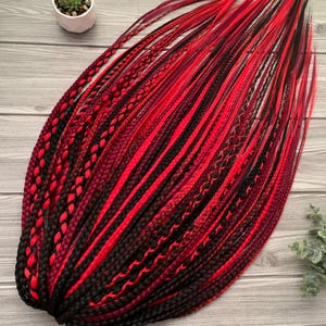 May include: A set of synthetic hair extensions featuring a mix of vibrant red, deep burgundy, and solid black strands. The collection includes a variety of textures, combining smooth straight hair with intricate three-strand braids and twists.