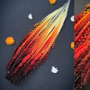 Peut inclure: Ensemble de dreadlocks colorés aux tons rouges, oranges, jaunes et noirs, disposés sur un fond sombre. Les dreadlocks présentent diverses textures et motifs, avec un effet dégradé. L'image est ornée de petites fleurs blanches et oranges.