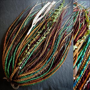 Może przedstawiać: Kolekcja ręcznie robionych dredów w różnych odcieniach brązu, zieleni i złota, z akcentami białej koronki i sztucznej zieleni. Plecione i skręcone tekstury tworzą estetykę boho. Dready ułożone są na ciemnoszarej powierzchni.