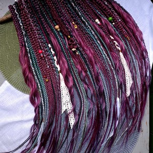 May include: A set of synthetic hair extensions featuring a mix of deep burgundy, dark purple, and muted teal braided strands. The hair is adorned with small wooden and metallic beads, thin white lace ribbons, and loose wavy hair sections.