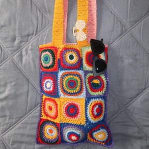 Puede incluir: Una bolsa de mano de crochet colorida con una correa rosa y amarilla. La bolsa está hecha de cuadrados con círculos de diferentes colores en el centro. Unas gafas de sol están metidas en la parte superior de la bolsa.