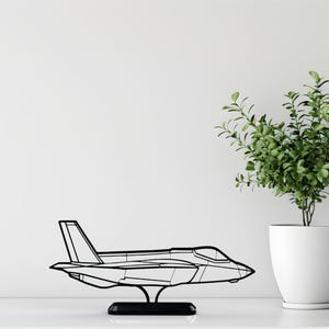 May include: A black metal airplane sculpture on a black base. The sculpture is a detailed outline of a jet fighter. A potted plant with green leaves sits to the right of the sculpture.