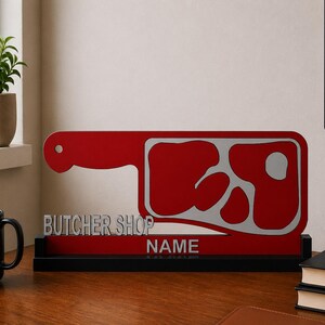 Op de afbeelding: Een rood en zilverkleurig bord in de vorm van een slagersmes met de tekst BUTCHER SHOP en NAME in zilveren letters. Het bord is gemonteerd op een zwarte rechthoekige voet, ontworpen om op een vlakke ondergrond te worden geplaatst.