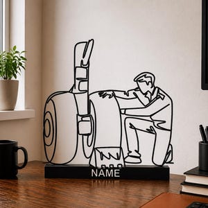 May include: A black metal wire art sculpture depicting a person kneeling beside a large aircraft landing gear assembly. The sculpture is mounted on a rectangular black base with the word NAME printed in white capital letters on the front.