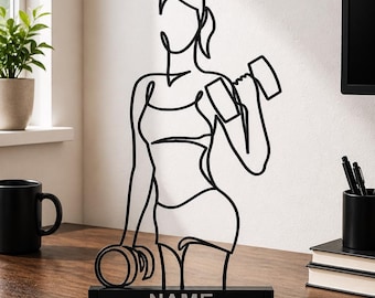 Peut inclure: Une sculpture en fil de métal noir représentant une personne soulevant des haltères. La silhouette est réalisée dans un style de ligne continue, montrant une personne en tenue de sport. La sculpture est fixée sur un socle noir avec le mot NAME.