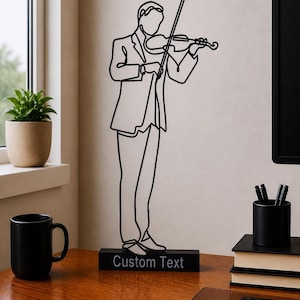 May include: A black metal sculpture of a violinist playing a violin, mounted on a black rectangular base. The base has the text "Custom Text". The sculpture is on a wooden desk with a black mug, a potted plant, and books.