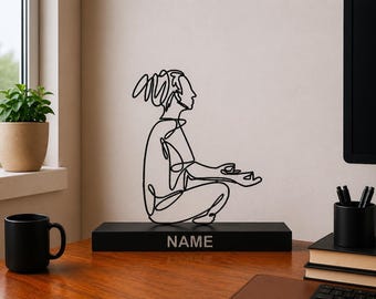 Peut inclure: Une sculpture en fil de métal noir représentant une personne en position de méditation assise avec les mains en mudra. La sculpture est fixée sur une base rectangulaire noire portant le mot NAME imprimé en police sans-serif gris clair.
