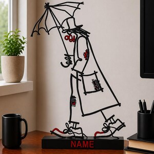 May include: A black wire sculpture of a figure wearing a hat and long coat, holding an umbrella. The figure features red accents on the shoes, patches, and face. It stands on a rectangular black base with the word NAME printed in red letters.