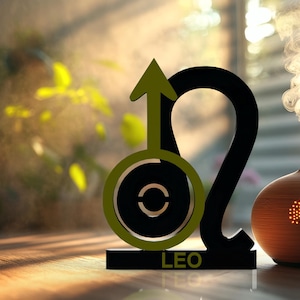 May include: A black and olive green Leo zodiac sign sculpture featuring a stylized lion symbol with an upward-pointing arrow. The base of the sculpture is black and displays the text LEO in olive green lettering, positioned next to a round diffuser.