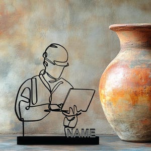 Puede incluir: Una escultura de metal negro de una persona con casco y sosteniendo una computadora portátil, con la palabra "NAME" en la base. A la derecha, un jarrón grande, texturizado, naranja y marrón con asa.