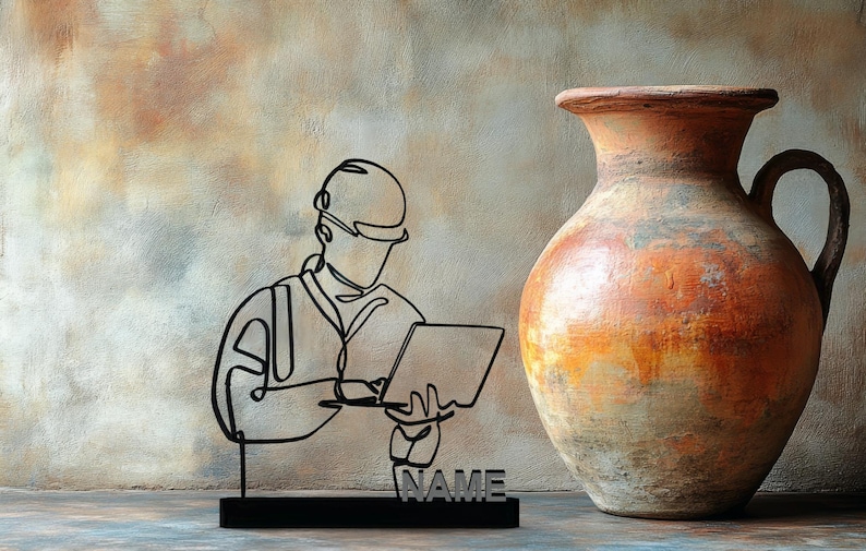 Puede incluir: Una escultura de metal negro de una persona con casco y sosteniendo una computadora port&aacute;til, con la palabra "NAME" en la base. A la derecha, un jarr&oacute;n grande, texturizado, naranja y marr&oacute;n con asa.