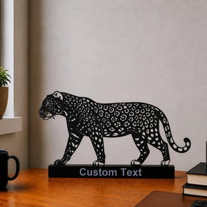 May include: Black metal jaguar sculpture with white spots, on a black base with "Custom Text". The sculpture is on a wooden desk, with a black mug, a potted plant, and a pen holder. The desk is near a window.