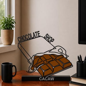 Könnte beinhalten: Eine schwarze Metalldrahtskulptur einer teilweise ausgepackten Schokoladentafel. Die Skulptur zeigt den Schriftzug "CHOCOLATE SHOP" aus schwarzem Draht und das Wort "CACAW" in weißem Druck auf dem rechteckigen schwarzen Sockel.
