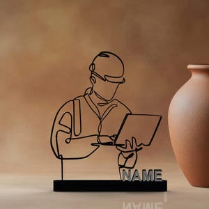 Puede incluir: Escultura de metal negro de una persona con casco trabajando en un port&aacute;til. La escultura est&aacute; sobre una base negra con la palabra "NAME" en letras plateadas. Un jarr&oacute;n marr&oacute;n est&aacute; a la derecha.