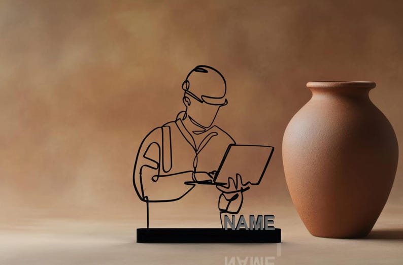 Puede incluir: Escultura de metal negro de una persona con casco trabajando en un port&aacute;til. La escultura est&aacute; sobre una base negra con la palabra "NAME" en letras plateadas. Un jarr&oacute;n marr&oacute;n est&aacute; a la derecha.