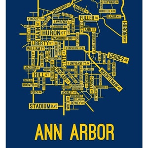May include: A yellow and blue map of Ann Arbor, Michigan, with the city's name in large yellow letters at the bottom. The map shows the major streets and avenues in the city, including Main Street, Huron Street, Liberty Street, and Geddes Avenue.