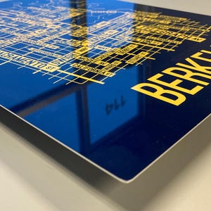May include: A blue metal print with a yellow map of Berkeley, California. The map includes street names and landmarks. The text "BERKELEY" is printed in large yellow letters at the bottom right corner.