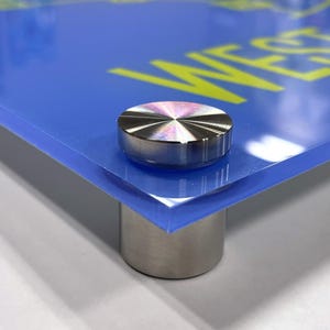 May include: Close-up of a blue acrylic print with a yellow inscription. The print is mounted on a clear acrylic stand with a silver metal base.