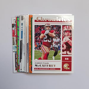 May include: A stack of football trading cards featuring a player in a red and gold uniform. The card on top has the word "Chronicles" at the top and the player's name, Christian McCaffrey, at the bottom. The cards are in a protective sleeve.