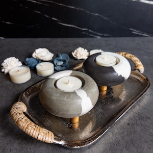 May include: Two gray and white concrete candle holders with wooden legs, each holding a white tea light candle. The candle holders are on a silver tray with a woven wicker handle.