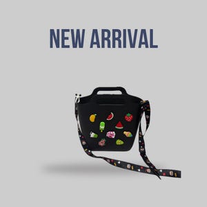 May include: Black tote bag with a handle and a detachable strap, decorated with colourful fruit-themed embellishments. The bag has the text "NEW ARRIVAL" in blue. The strap features a repeating pattern of small cartoon characters.