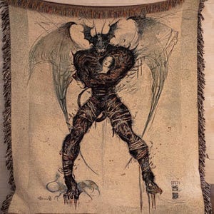 May include: A tapestry wall hanging featuring a black and white illustration of a winged demon holding a woman. The demon has horns and a long tail. The tapestry has a beige background with a brown border.