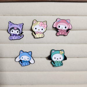 May include: Five enamel pins featuring cartoon characters dressed in costumes. The characters are a purple cat, a yellow cat, a pink rabbit, a blue dog, and a green dog. Each pin has a different costume and a different color scheme.