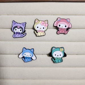 May include: Five enamel pins featuring different cartoon characters. The pins are in the shape of cats and feature different colors and designs. The characters are wearing different outfits and accessories.