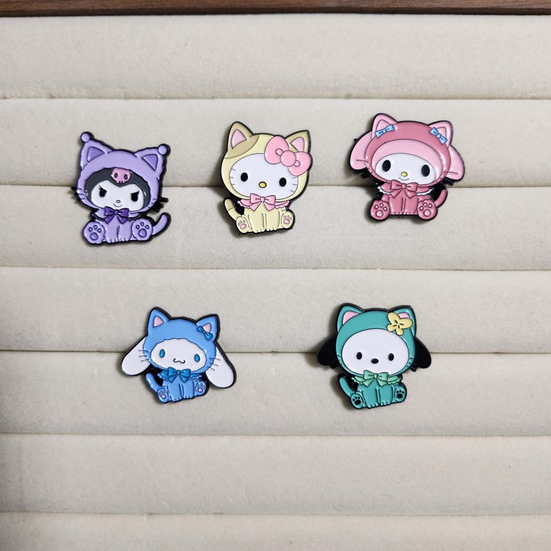 May include: Five enamel pins featuring different cartoon characters. The pins are in the shape of cats and feature different colors and designs. The characters are wearing different outfits and accessories.