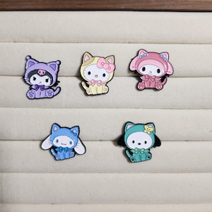 May include: Five enamel pins featuring cartoon characters wearing different colored hooded outfits. The characters are a cat, a rabbit, and a bear. The pins are all in different poses and have different expressions on their faces.